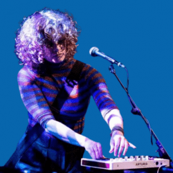 A musician with curly hair performing on stage, playing a small keyboard with a microphone in front of her. She wears a striped top and stands against a blue backdrop, focused on her performance.