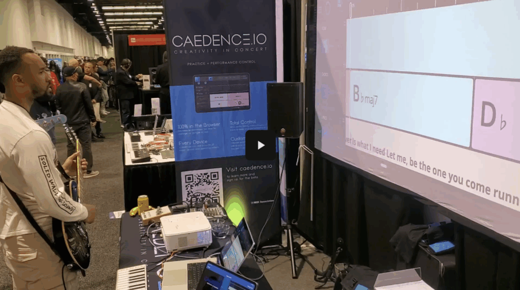 A man plays electric guitar at a booth with a blue CAENDENCE.IO banner. A projector displays chords and lyrics on a screen. Laptops, cables, and audio equipment are on the table. People walk in the background at an indoor event.