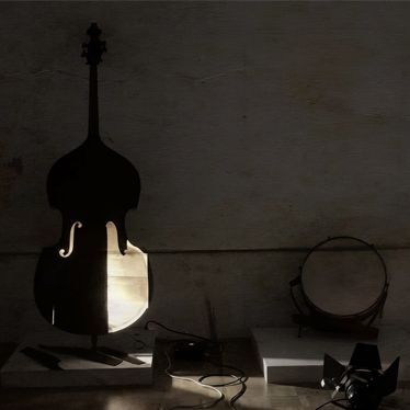 A double bass and a tambourine are displayed on separate platforms in a dimly lit room. Light shines through the double bass, casting shadows, with a spotlight and power cords nearby.