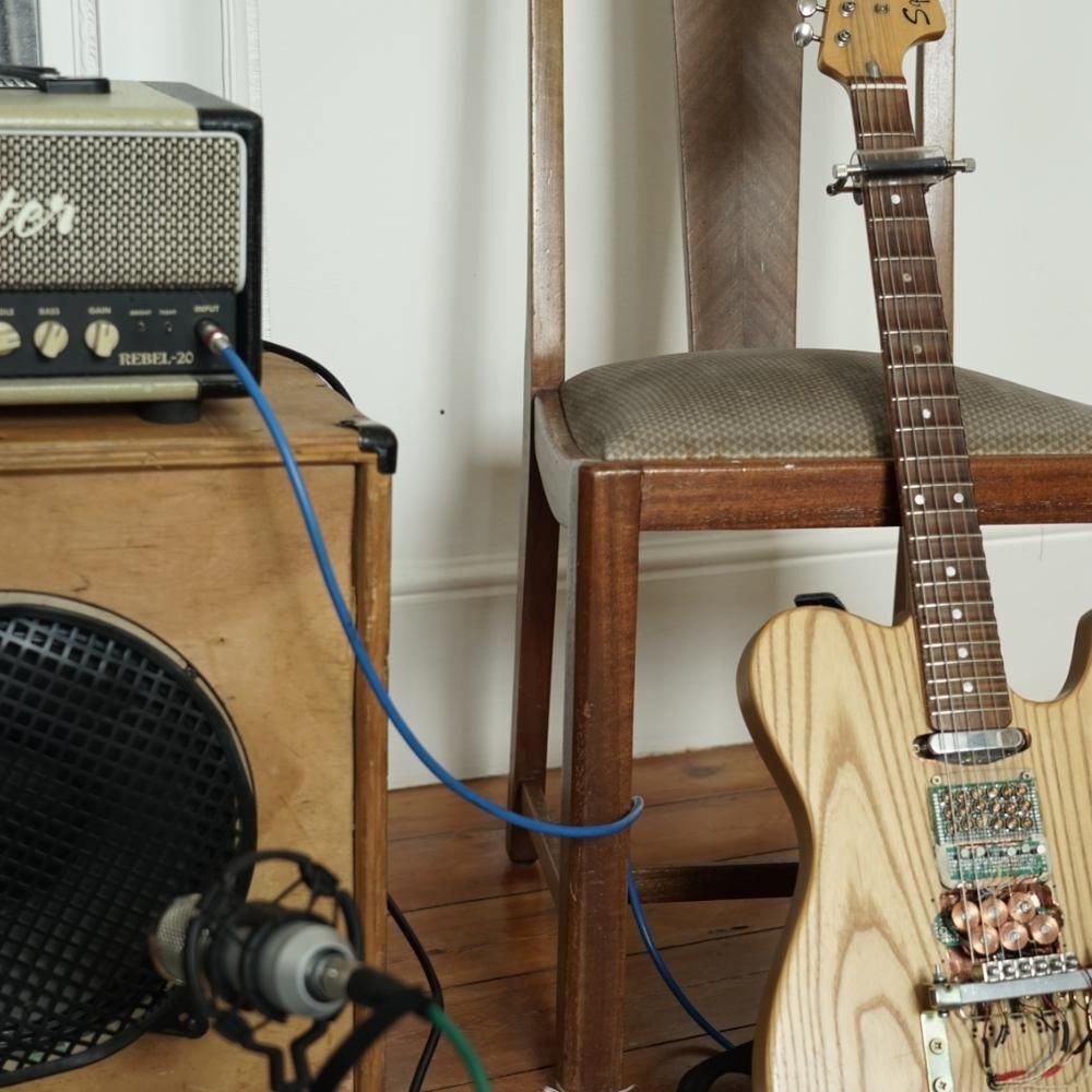 A light wood electric guitar leans against a padded chair next to a small guitar amplifier and speaker on a wooden floor, with a microphone positioned in front.
