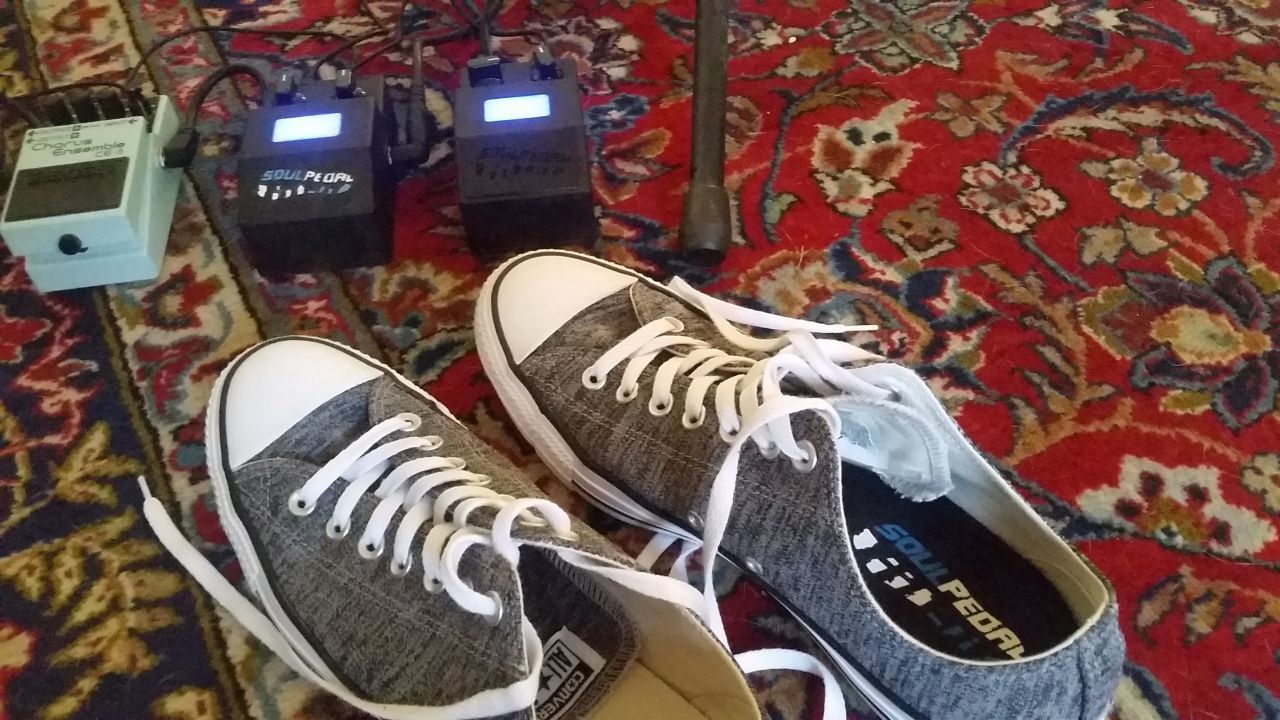 A pair of gray sneakers with white laces are placed on a colorful patterned rug near three guitar effect pedals and a microphone stand.
