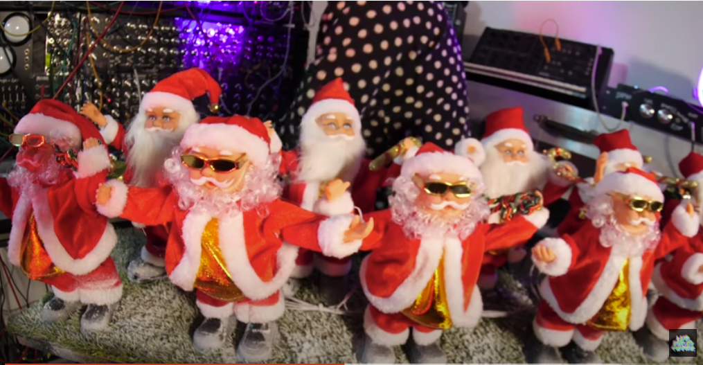 Eight toy Santa Claus figures wear sunglasses and festive red outfits, standing on a sparkly surface with electronics, lights, and wires in the background. A person in polka-dot clothing is partly visible behind them.