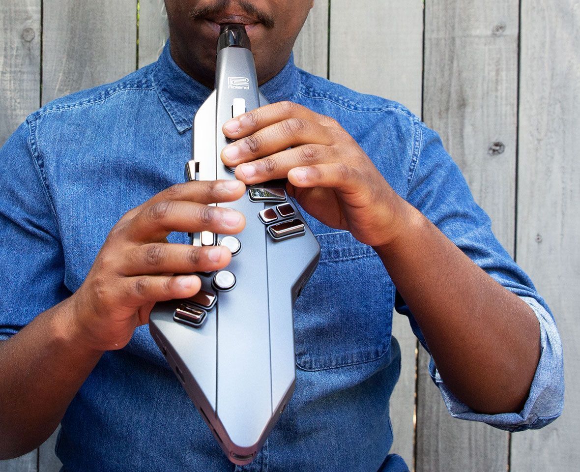 A person in a blue denim shirt plays an electronic wind instrument in front of a wooden fence, using both hands to press buttons on the device.