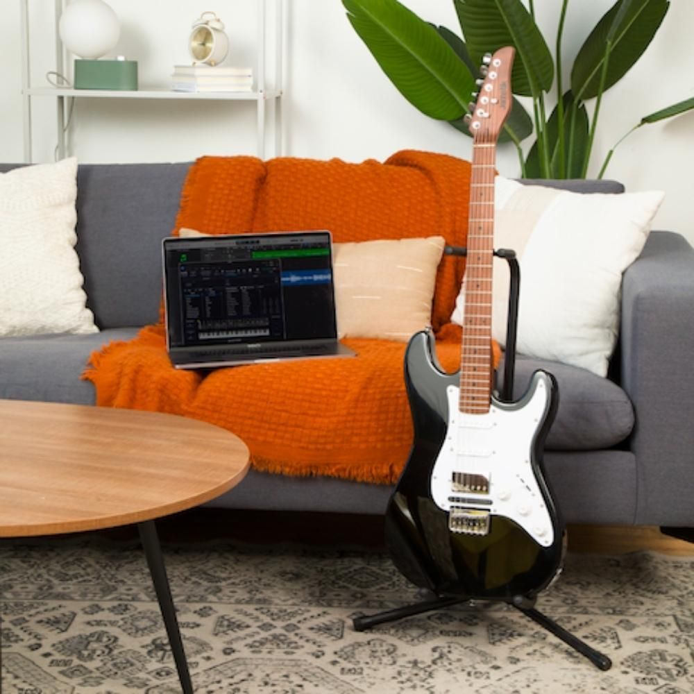 A black electric guitar on a stand sits next to a gray couch with orange and white pillows and a cozy orange blanket. An open laptop is on the couch, and a green plant is in the background.