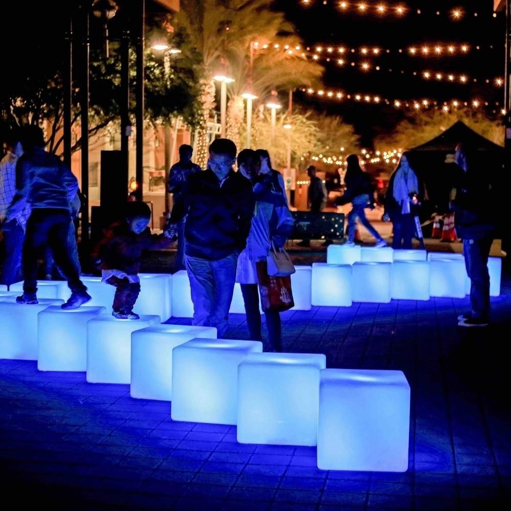 People walk and play on glowing blue light cubes outdoors at night, with string lights above and trees in the background, creating a festive atmosphere.