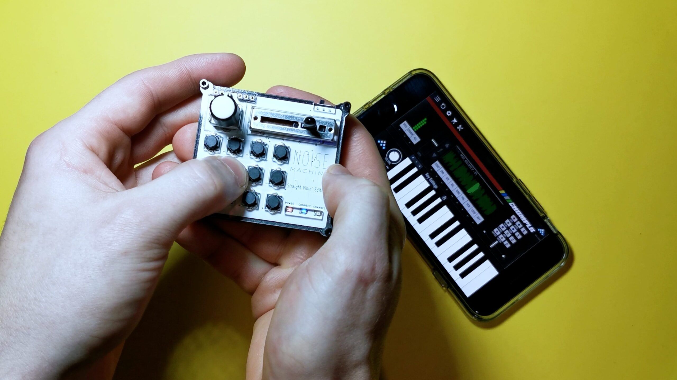 Two hands holding a small music synthesizer with knobs and buttons, next to a smartphone displaying a piano keyboard app, all on a yellow background.