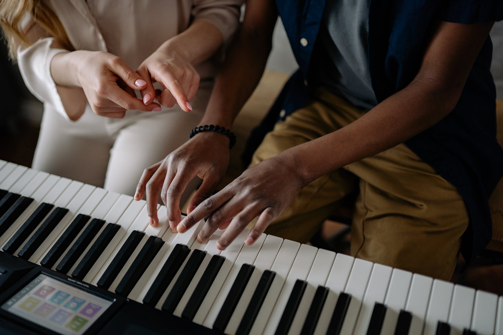 Two people sit at an electric piano; one person is playing the keys while the other person points and guides them, suggesting a piano lesson or instruction. Only their hands and part of their laps are visible.