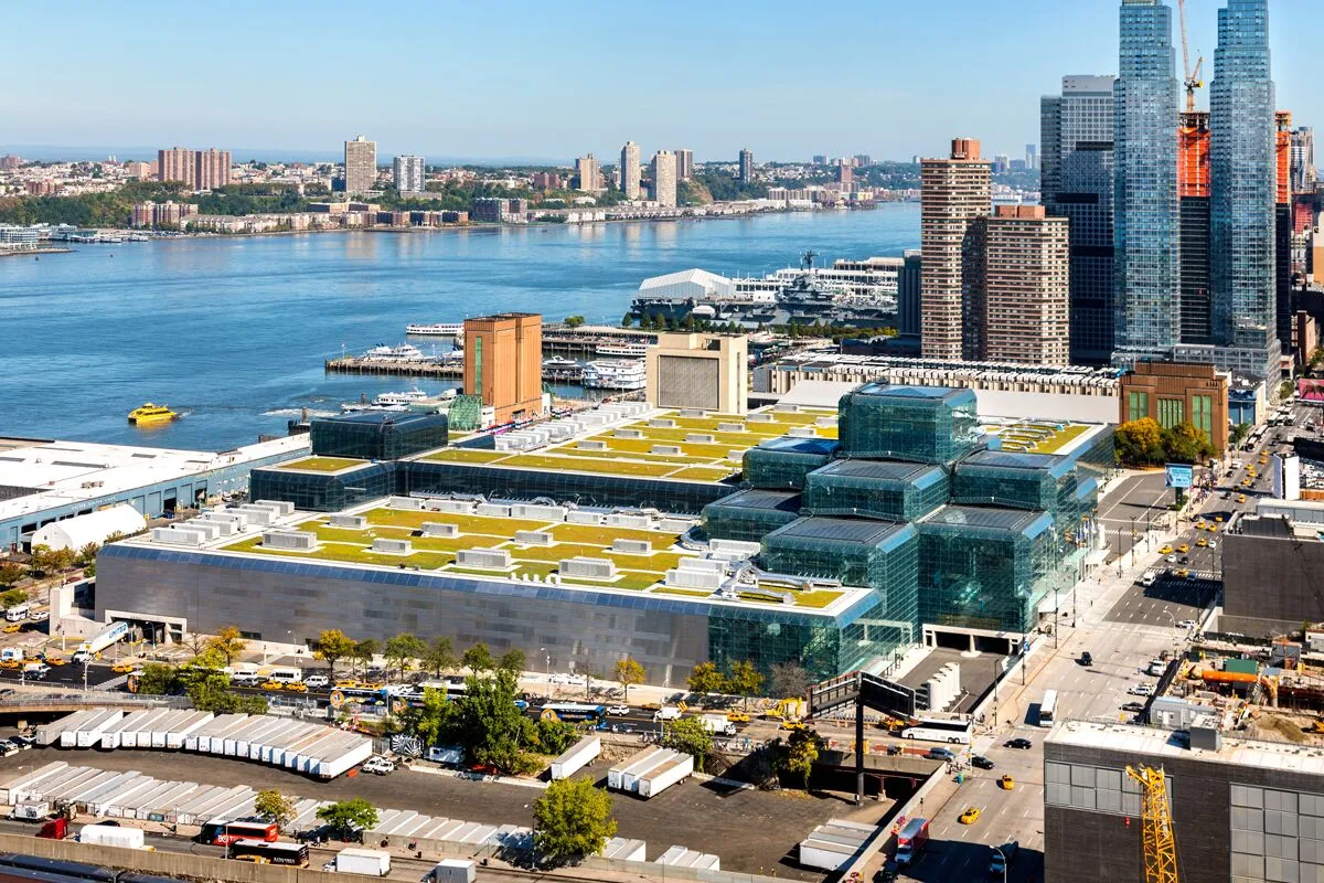Aerial view of the Javits Center with its glass roof and green spaces, located along the Hudson River in Manhattan, New York City, with skyscrapers and waterfront visible in the background.
