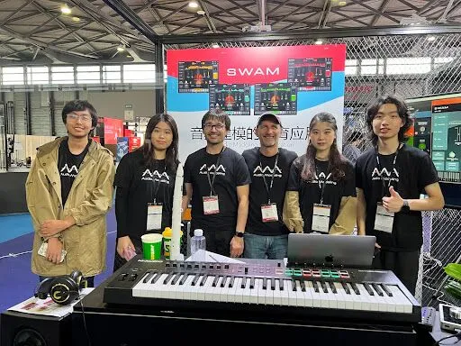 Six people stand together at a booth with a SWAM sign, wearing matching black shirts, behind a keyboard and audio equipment at an indoor event or exhibition.