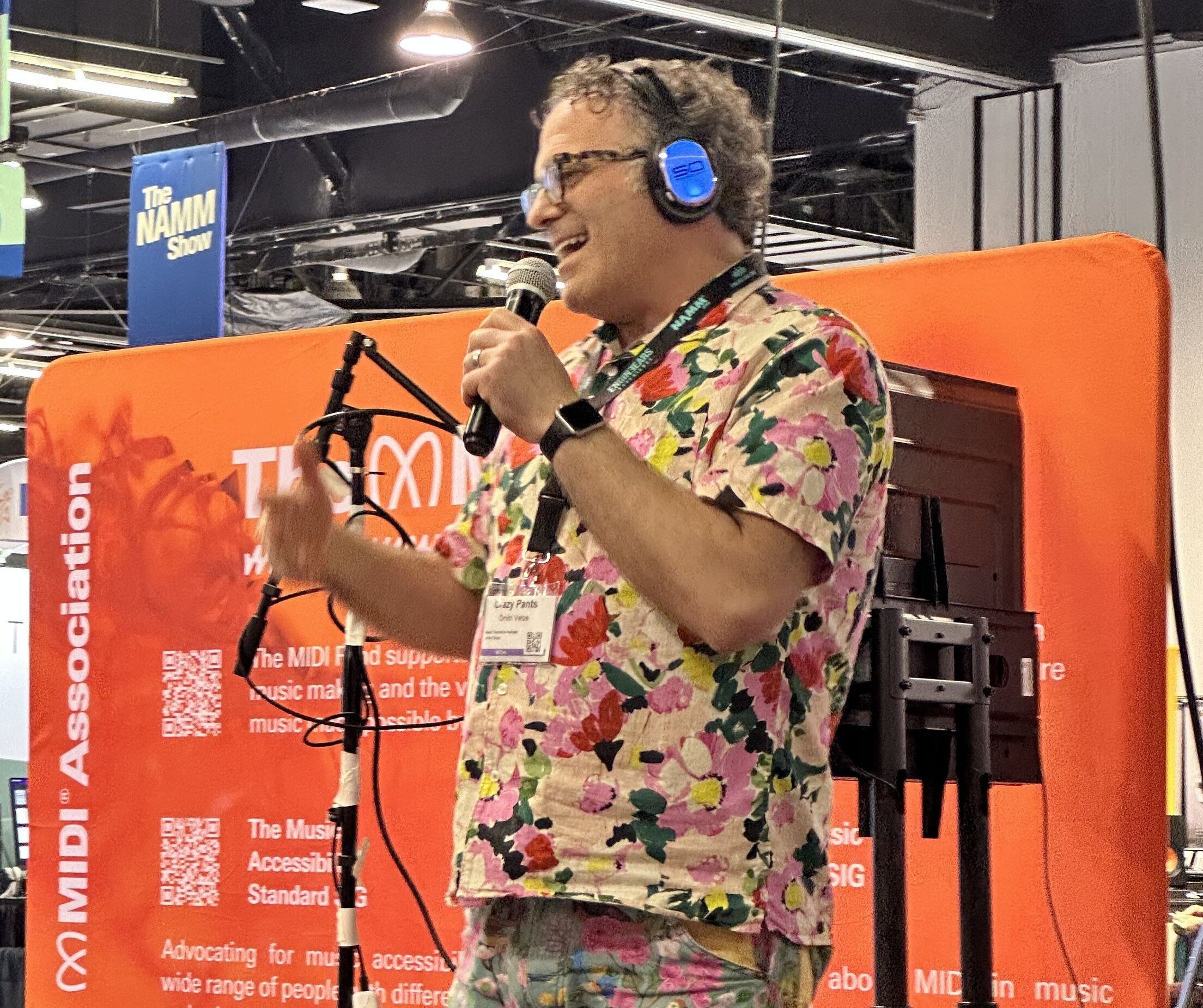 A person wearing blue headphones and a colorful floral shirt speaks into a microphone in front of a red MIDI Association display at an indoor event.