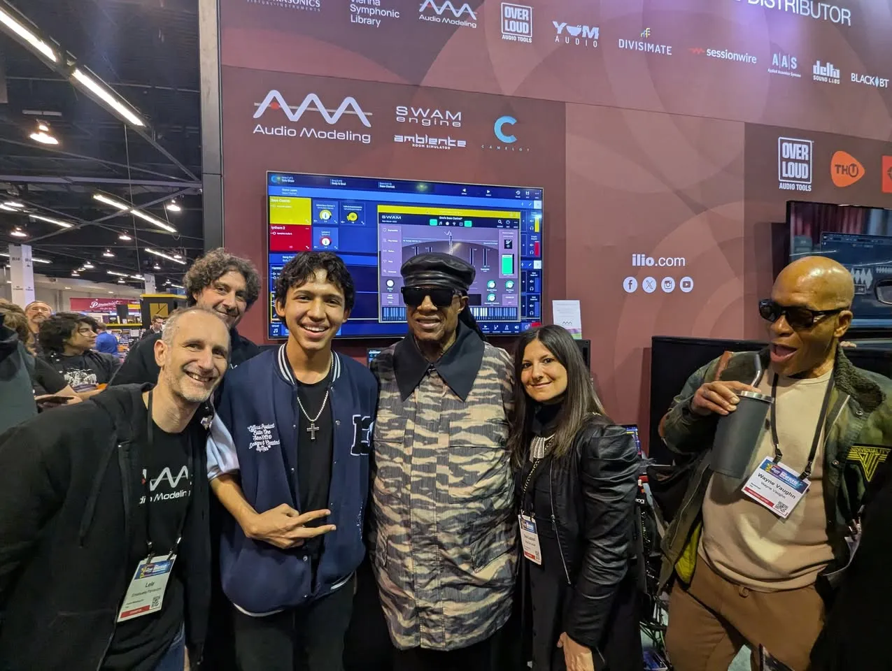A group of six people smile and pose together at a music technology event, standing in front of a booth with audio software displays and banners. One person wears sunglasses and a patterned jacket.
