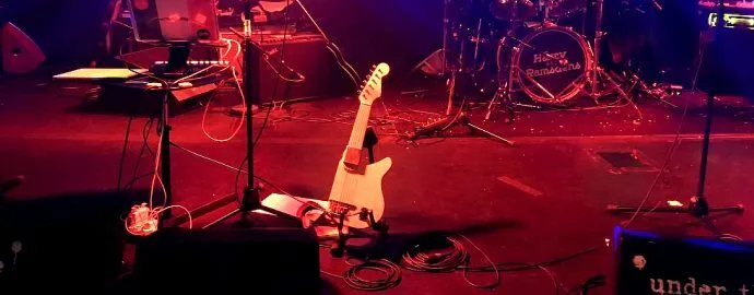 A dimly lit stage with a guitar resting on a stand, surrounded by cables, microphones, a drum set, and music equipment, ready for a live music performance.