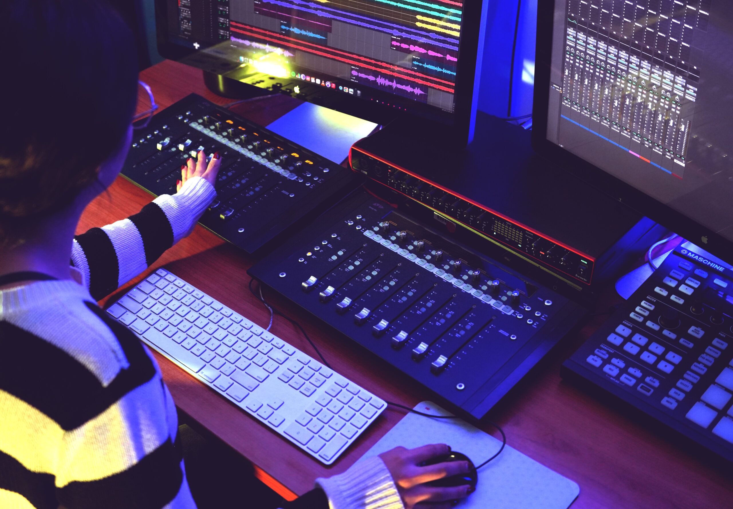 A person working at a music production studio desk with audio mixing equipment, a computer keyboard, and multiple monitors displaying colorful audio tracks and mixing software.