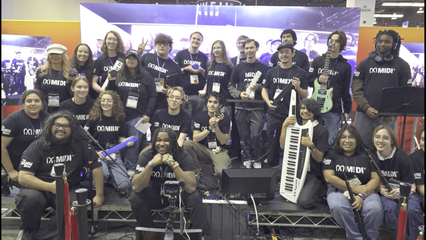 A large group of young people wearing matching black “MIDI” shirts pose together on a stage with musical instruments, including guitars and a keytar. Everyone is smiling and looking at the camera.