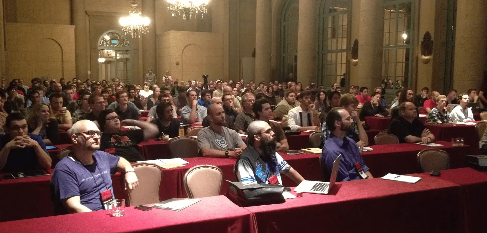 A large audience of people sits attentively at round tables covered with red tablecloths in an ornate, chandelier-lit conference room with tall arches and windows.
