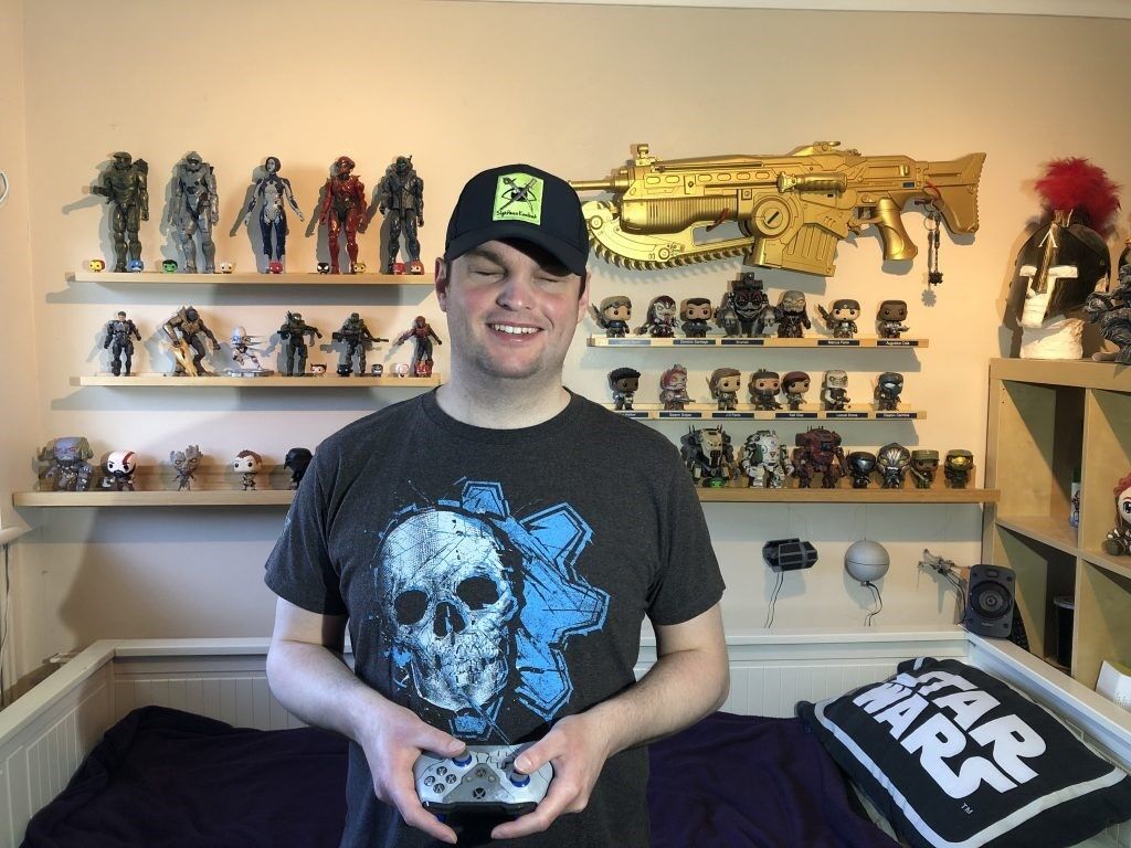 A smiling person wearing a black cap and graphic t-shirt stands in a bedroom holding a game controller. Shelves behind them display action figures and collectibles, including a large gold gun and a Star Wars pillow on the bed.