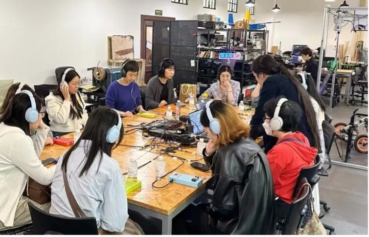 A group of people wearing headphones sit around a large table covered with audio equipment and laptops, engaged in discussion in a modern, well-lit workspace.