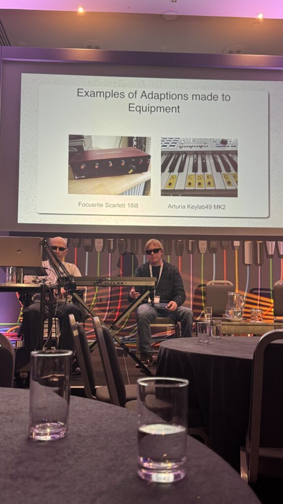 Sam Prouse and David Shervill sit on a stage with microphones, in front of a screen showing examples of equipment adaptations: a Focusrite Scarlett 18i8 and an Arturia Keylab49 MK2. The background features colorful stripes.