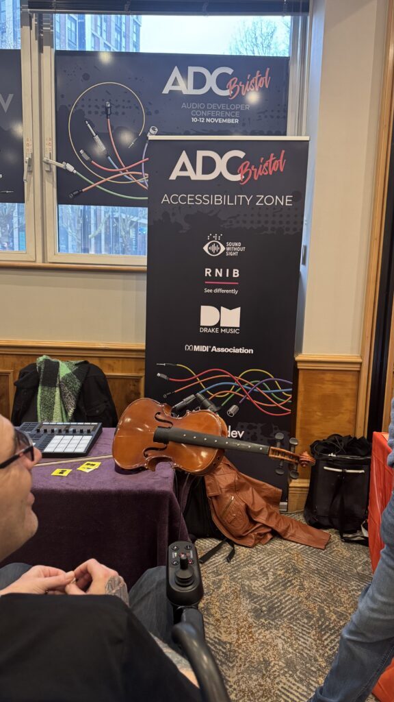 A cello rests on a table in front of a banner reading ADC Bristol Accessibility Zone at an indoor event. Various accessibility organizations’ logos appear on the banner. People are visible in the foreground.