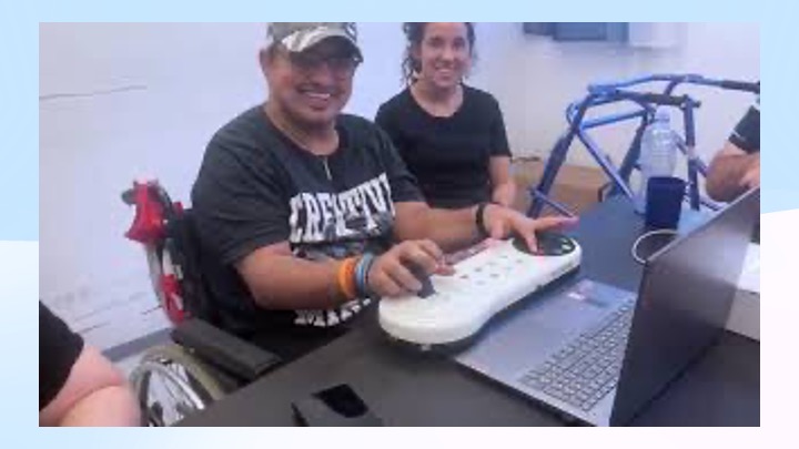 A smiling man in a wheelchair uses an adaptive controller connected to a laptop, with a woman sitting beside him. A blue mobility aid and water bottle are visible in the background.