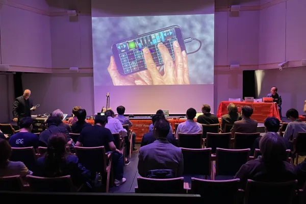 Audience seated in a dimly lit auditorium watches a presentation featuring a large screen displaying a hand using a music app on a smartphone. Two presenters are at tables in the front of the room.