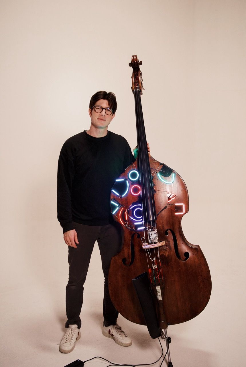 A person wearing glasses, a black sweater, and sneakers stands beside an upright bass decorated with colorful neon lights, against a plain light background.