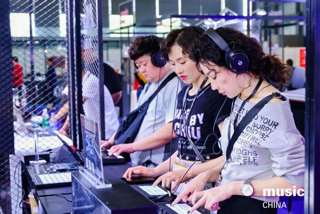 Three young people wearing headphones use electronic music equipment at a booth. They are focused on creating music, with laptops and controllers in front of them. The scene appears to be at a music expo or event.