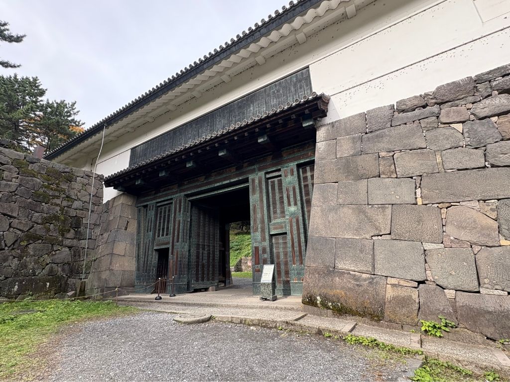 A large traditional Japanese wooden gate with metal details set in tall stone walls, surrounded by greenery and a gravel path, under a cloudy sky.