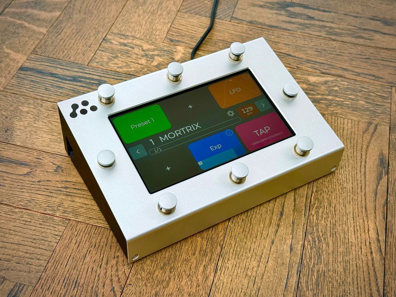 A rectangular electronic device with eight silver knobs and a touchscreen displaying colorful controls sits on a wooden floor. A black cord extends from the back.