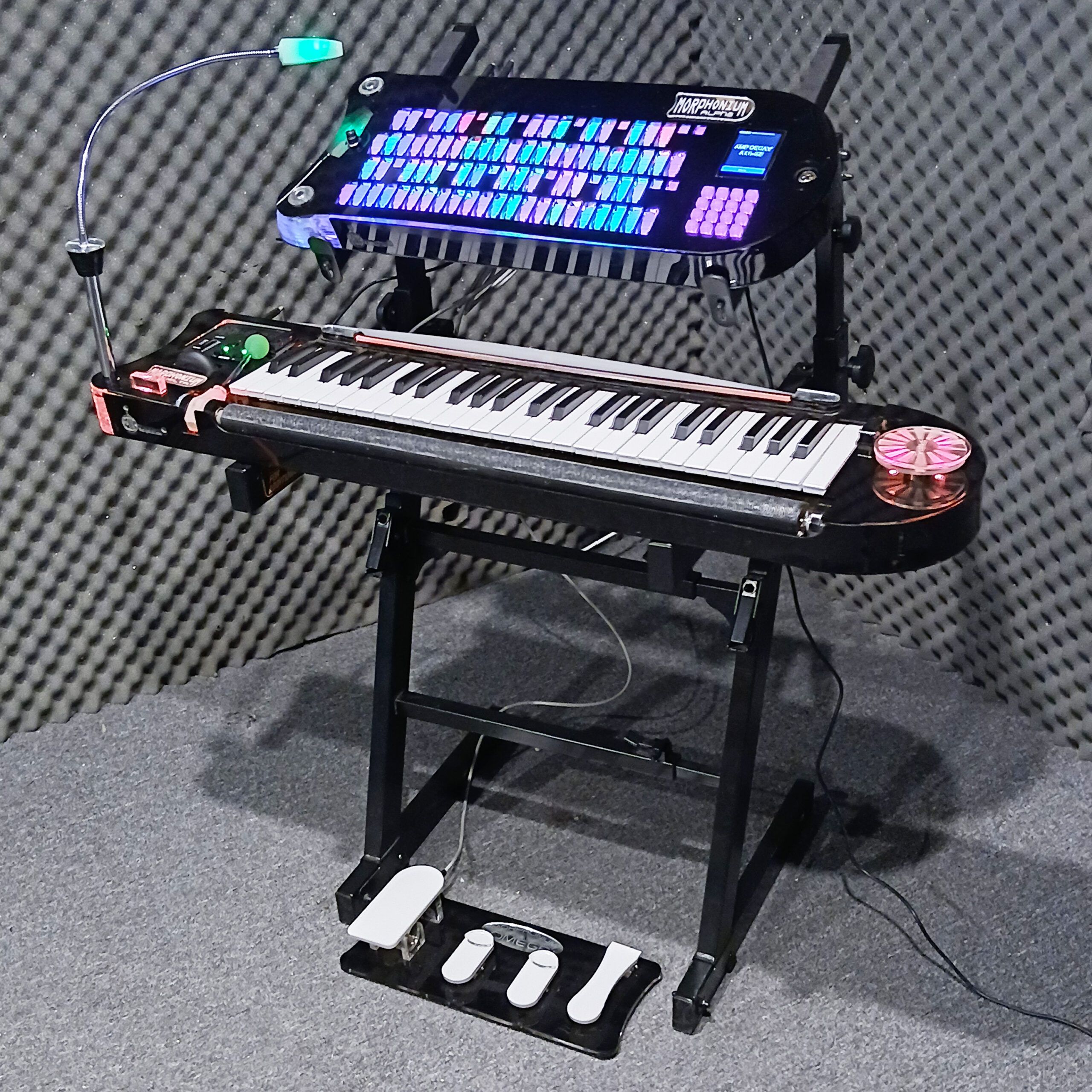 A futuristic electronic keyboard setup with illuminated controls, a touchscreen display, three foot pedals, and a small reading light, all arranged in a soundproofed room.