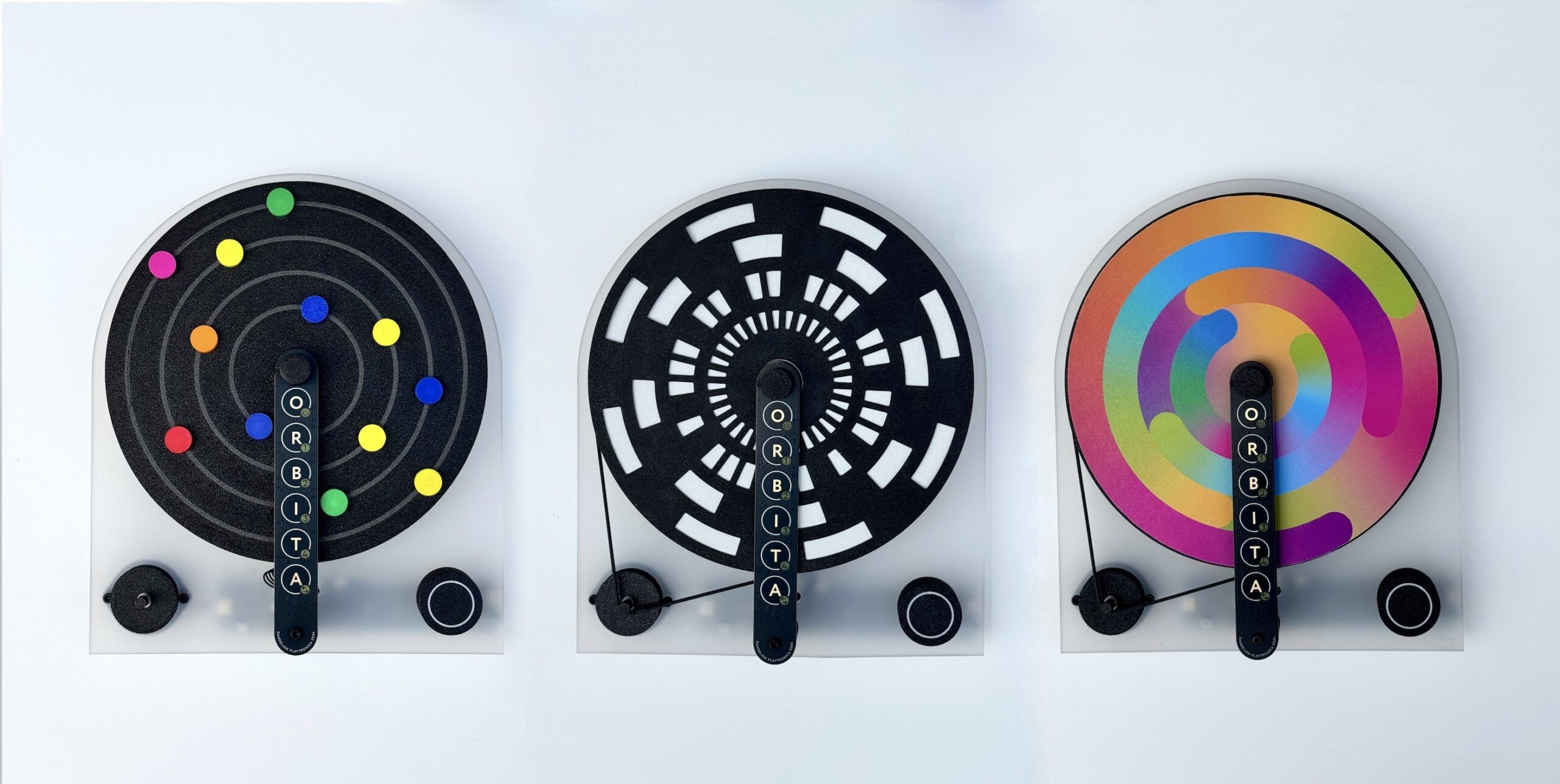 Three circular black-and-white and colorful abstract spinning discs are displayed side by side, each mounted on a clear stand labeled ORBITA with a black arm attached at the front.