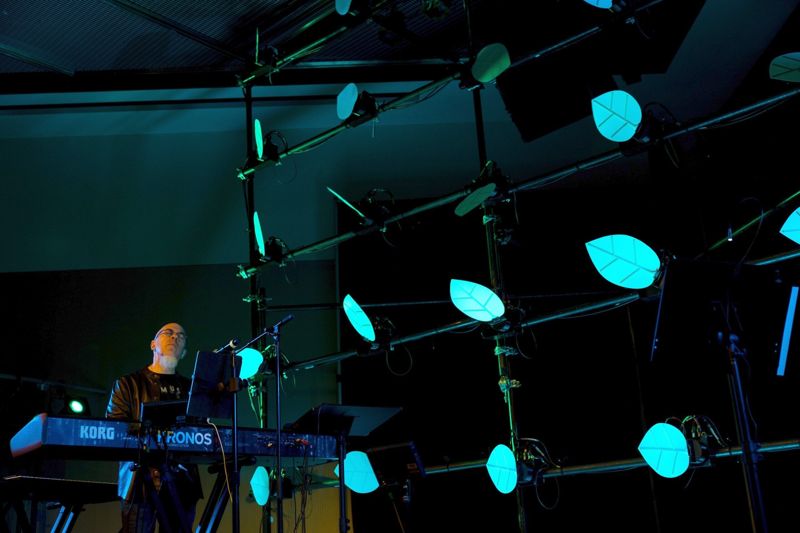A musician plays a Korg Kronos keyboard on stage, surrounded by an artistic installation of illuminated, leaf-shaped lights attached to a metal frame. The lighting is mostly blue and green, creating a futuristic atmosphere.