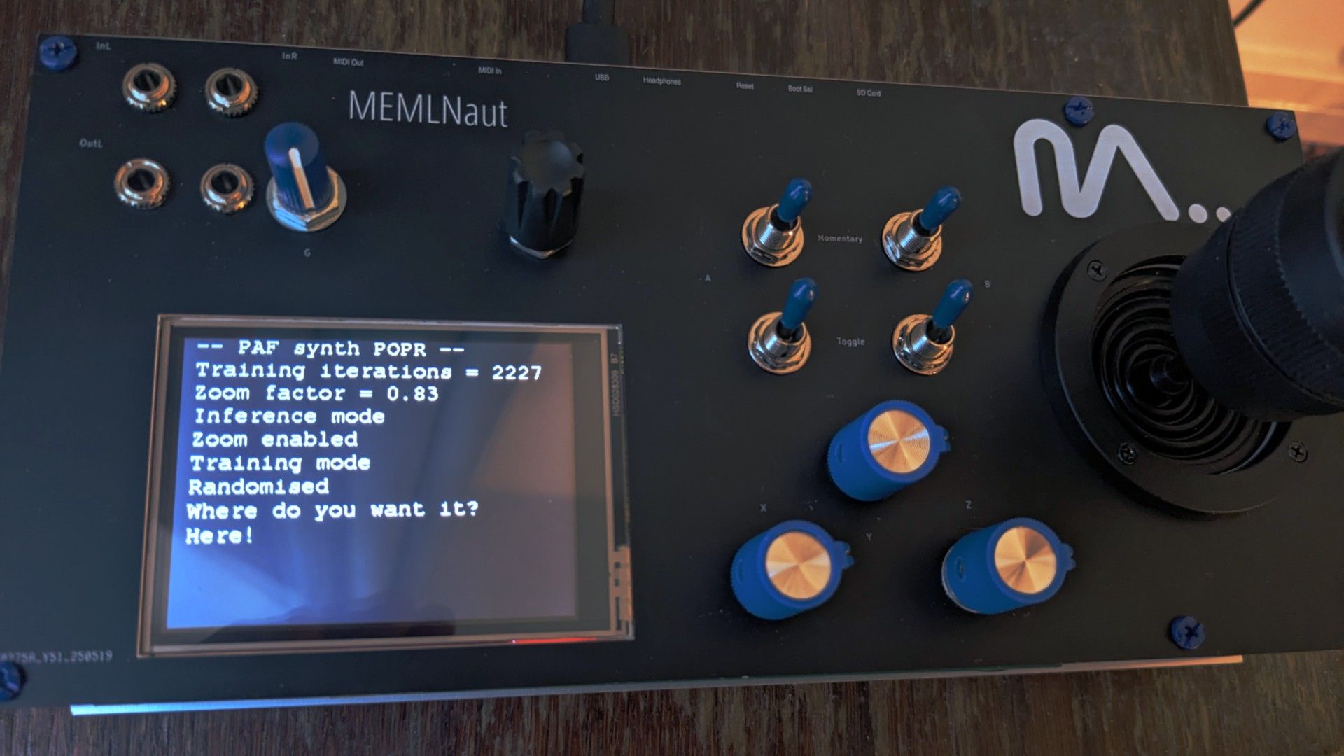 A close-up of a MEMLNAut device with a screen displaying machine learning settings, toggle switches, blue knobs, and a joystick on the right side. The screen shows training iterations and mode options.