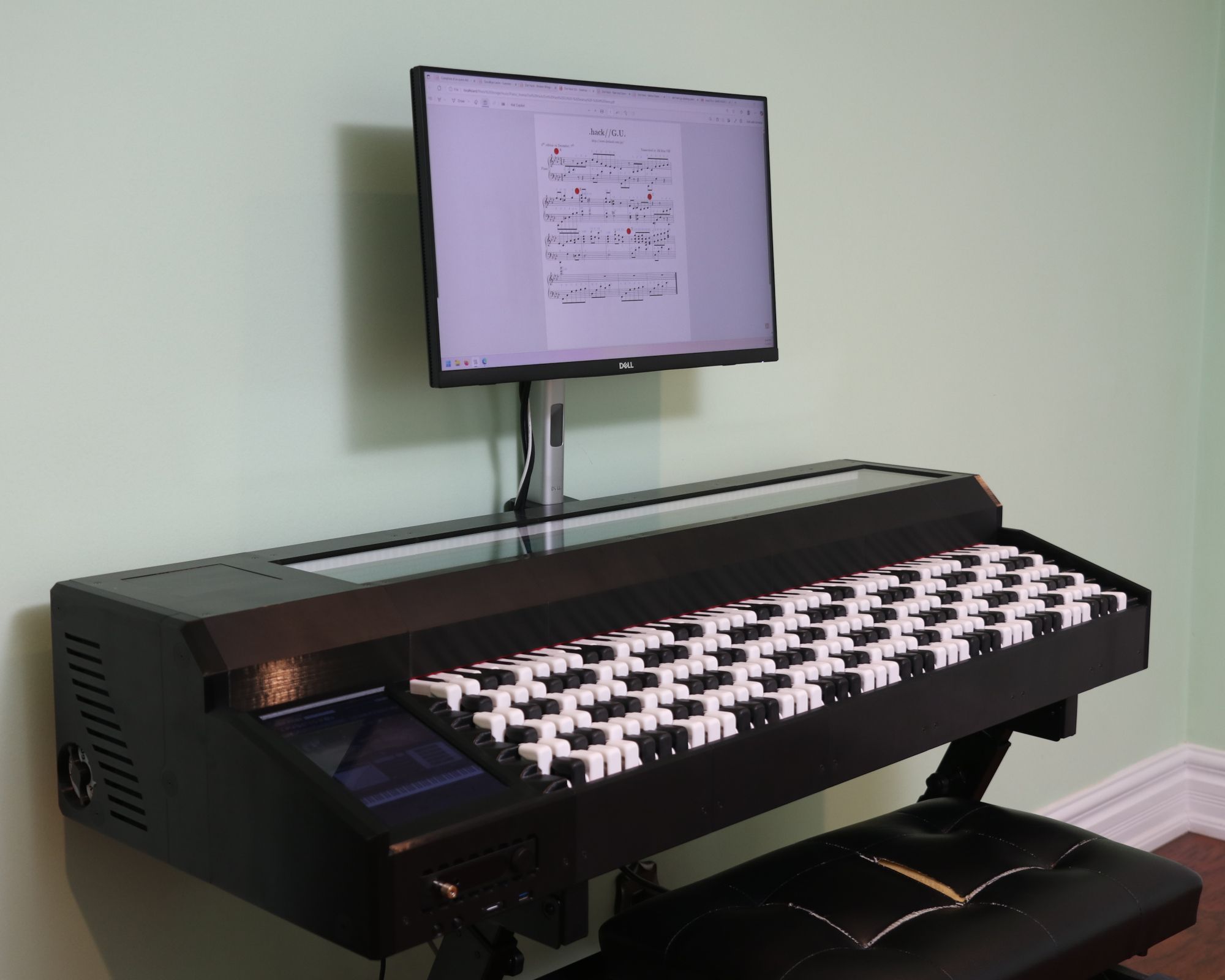 A modern electronic instrument with numerous white and black rectangular keys sits against a green wall, beneath a monitor displaying digital sheet music.