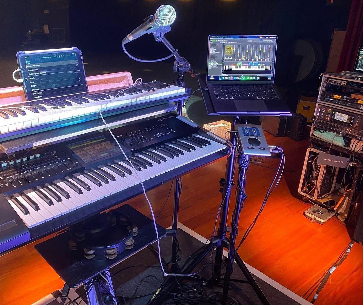 A music setup on stage featuring two stacked keyboards, a microphone, a laptop displaying audio software, a tablet, and various audio equipment and cables, all under warm lighting.