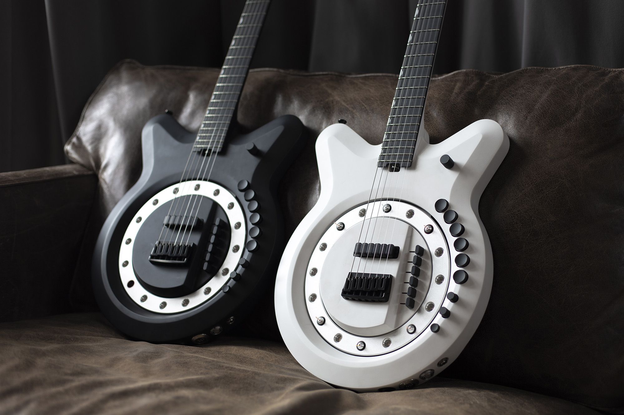Two modern electric guitars, one black and one white, sit side by side on a brown leather couch. Both have circular designs with multiple buttons and unique, futuristic features on their bodies.