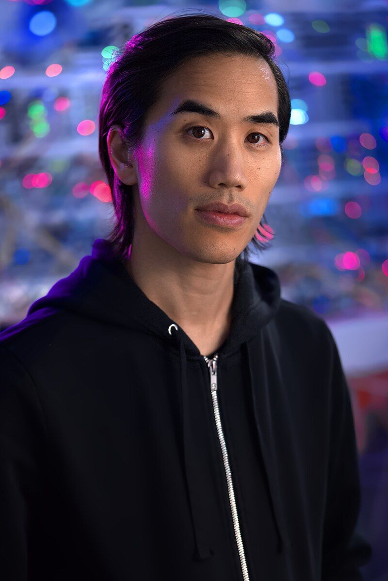 A person with straight black hair, wearing a black zip-up hoodie, stands in front of a colorful, blurred background with blue, pink, and green lights. The lighting casts a slight purple tint on their face.