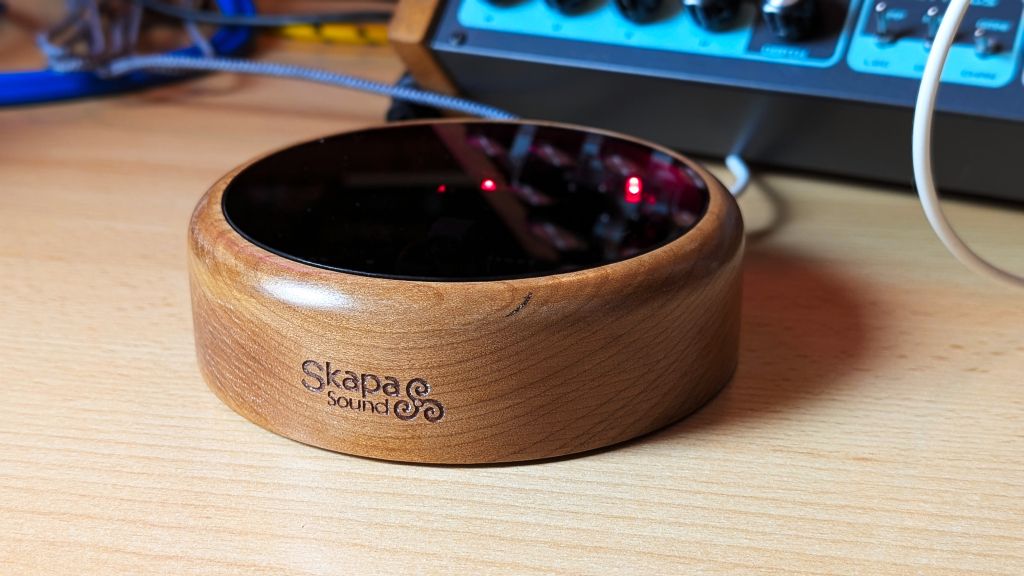 A wooden electronic device labeled Skapa Sound sits on a desk, surrounded by cables and audio equipment in the background. The device has a glossy black top.