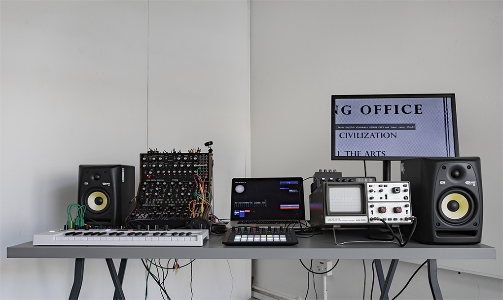 A music production setup on a gray table with speakers, a keyboard, a modular synthesizer, a laptop, an oscilloscope, a controller, and a monitor displaying text in a minimalist, white-walled room.