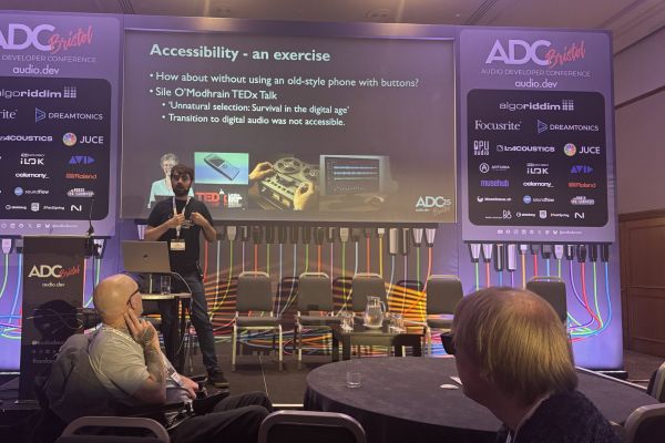 Jay Pocknell from SoundWithoutSight presents on stage at the ADC conference, discussing accessibility. A slide behind them references a TEDx Talk and features images of a vintage phone and retro equipment. Audience members watch and listen.