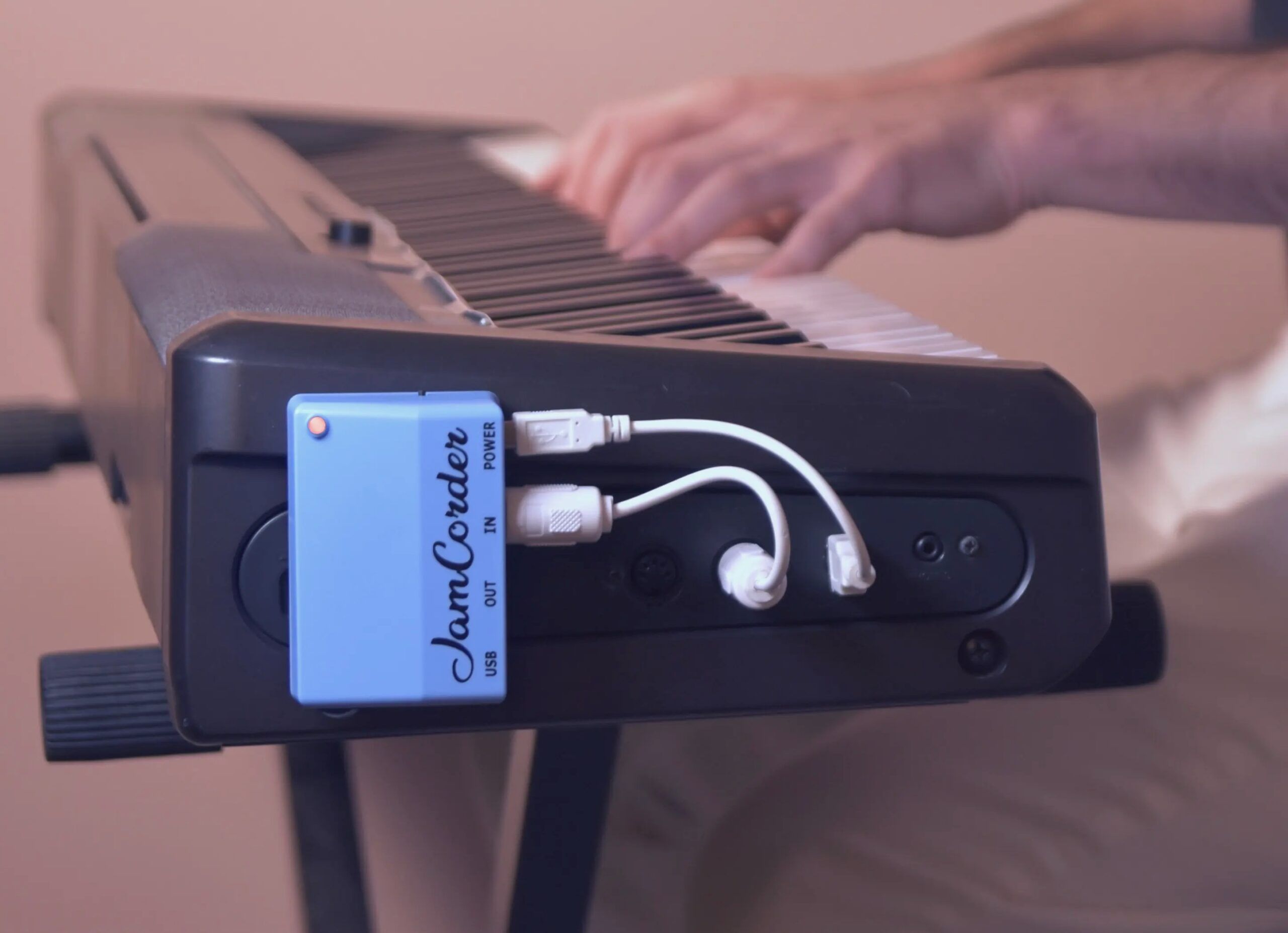 Close-up of a person playing an electronic keyboard, with a blue device labeled JamCorder connected to the keyboard via multiple white cables. The background is out of focus.