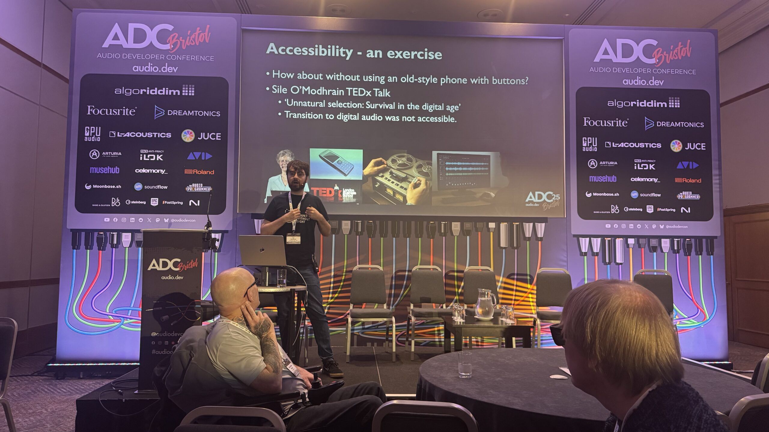 Jay Pocknell from SoundWithoutSight presents on stage at the ADC conference, discussing accessibility. A slide behind them references a TEDx Talk and features images of a vintage phone and retro equipment. Audience members watch and listen.