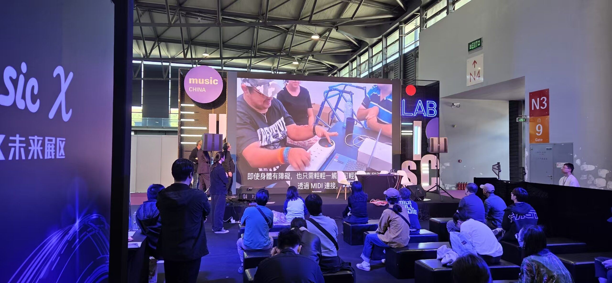 A group of people sit on benches facing a stage with a large screen displaying a demonstration of music equipment at an indoor event called music CHINA. Purple lighting and expo signs are visible in the background.