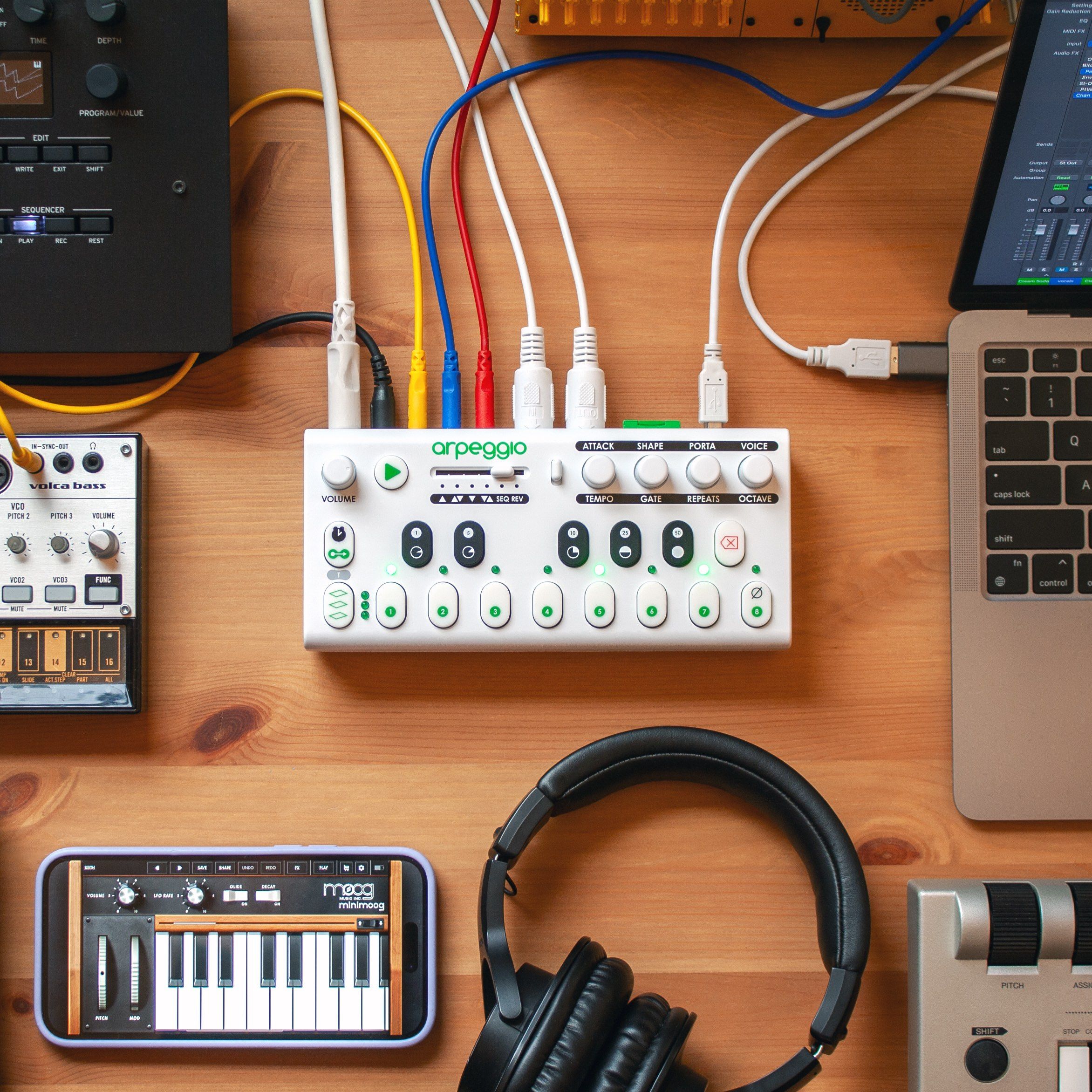 A tabletop music production setup with synthesizers, a MIDI controller labeled arpeggio, headphones, a laptop, various audio cables, and other electronic music equipment on a wooden desk.