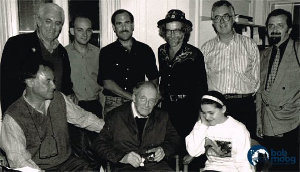 A group of nine people, including men and one young girl, posing together indoors. Some are standing and some are seated. They are smiling or looking at the camera in a casual group photo. A logo in the corner reads bob moog.