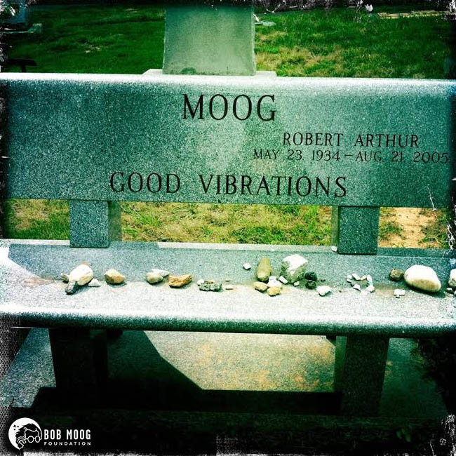 A stone bench engraved with “MOOG, Robert Arthur, May 23 1934 – Aug 21 2005, GOOD VIBRATIONS,” adorned with small stones on its seat. The Bob Moog Foundation logo is in the lower left corner.