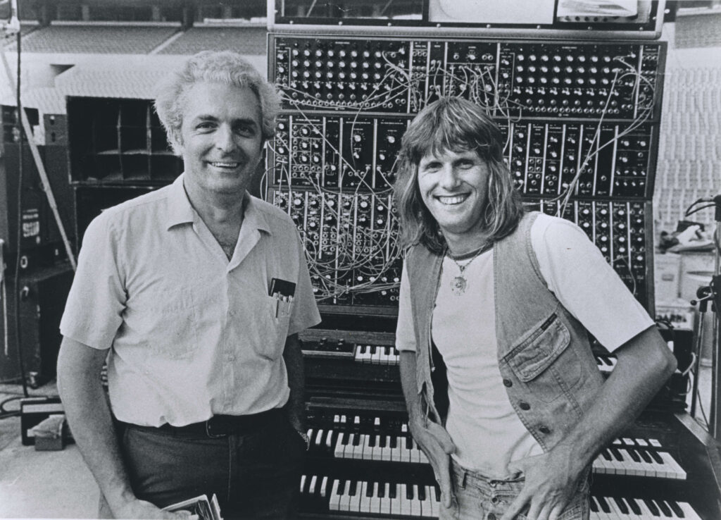 Two men stand smiling in front of a large vintage modular synthesizer and keyboards, with cables and dials visible, in what appears to be a music studio or stage setting.