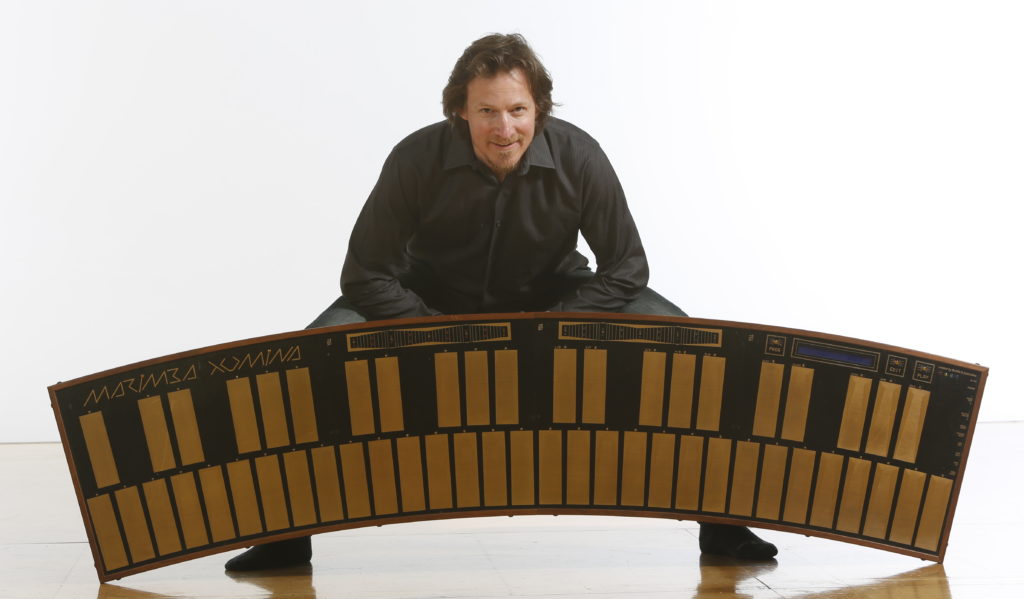 A man dressed in black crouches behind a large, curved electronic instrument called the Marimba Lumina, which has multiple rectangular pads and a display screen on its surface. The background is plain white.