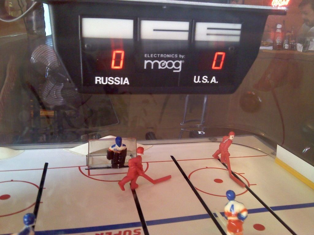 Tabletop hockey game showing teams Russia and U.S.A. with a digital scoreboard reading 0-0. Three red players and one blue-and-white player are on the rink, with a goalie in front of the net.
