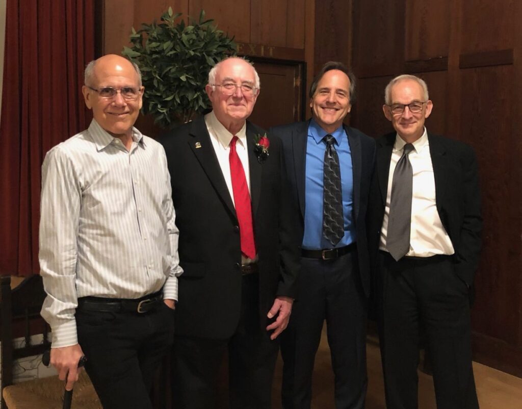 Dave Smith, Tom Oberheim, Marcus Ryle and Roger Linn in formal and semi-formal attire stand together indoors in front of wood-paneled walls and a potted plant. One man wears a red tie and boutonniere, while the others wear dress shirts or suits.